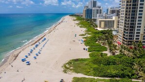 Ein Strand mit Reihen blauer Sonnenschirme, einem Sandufer und hohen Gebäuden im Hintergrund.