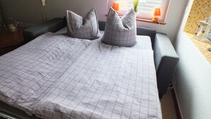Una cama bien hecha con colcha a cuadros y dos almohadas, una mesita de noche con lámpara, y una ventana con persianas y una planta.