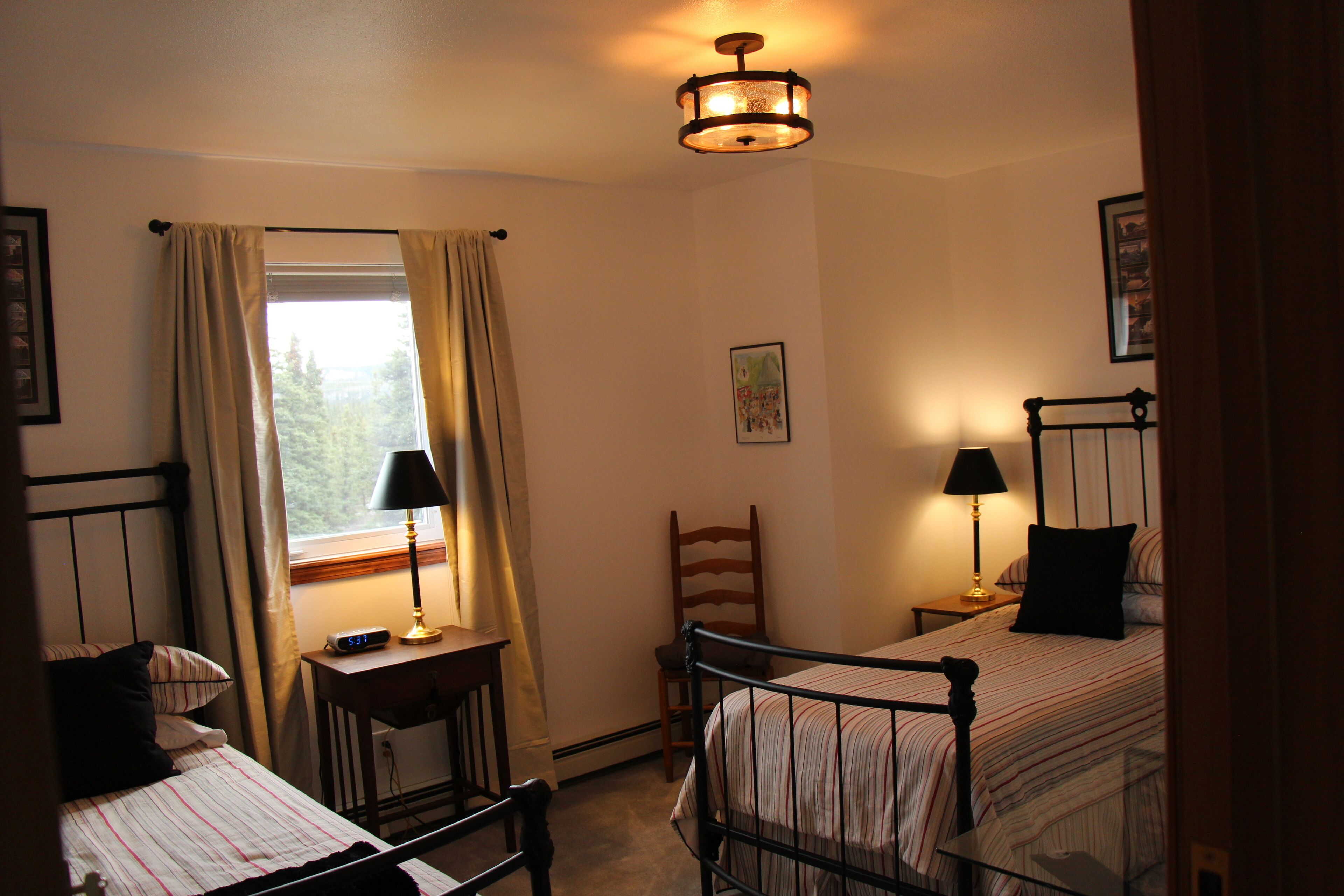 Ein Schlafzimmer mit zwei Betten, einem Fenster mit Vorhängen, einem Schreibtisch mit Lampe und einem Wandbild.