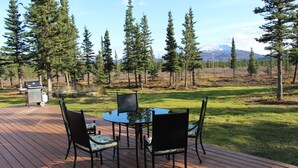 A deck with a glass-top table and chairs, surrounded by a forest and a mountain in the background.