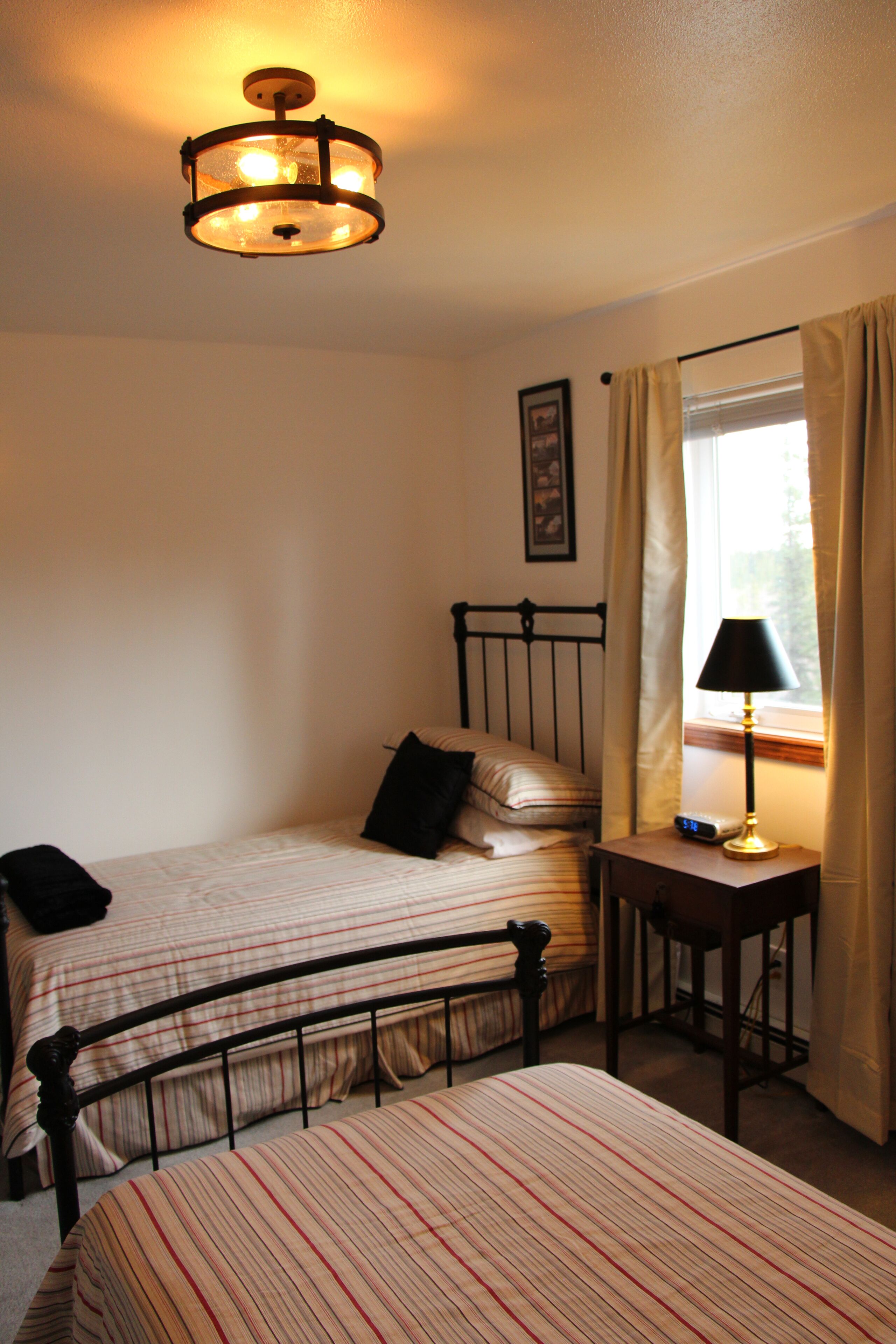 A bedroom with two single beds, a nightstand with a lamp, a window with curtains, and a ceiling light fixture.