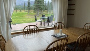 A dining area with a wooden table and chairs, a view of a grassy landscape with trees and mountains through large windows, and a patio area with outdoor furniture.