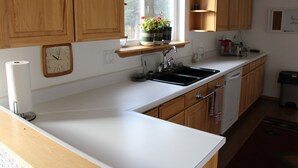 A kitchen with wooden cabinets, a white countertop, a sink, a clock, and a window with plants.