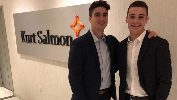 Deux hommes en costume, debout devant une enseigne sur laquelle on peut lire « Kurt Salmon ».