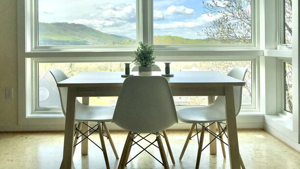 A dining area with a table and chairs, large windows with a view of hills and trees, and a potted plant on the windowsill. - Greensview Apartment | 2 Bedroom, 2 Bath - Beautiful views! (Charlottesville)