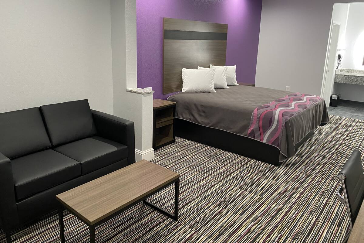 A modern hotel room with a bed, a black sofa, a wooden coffee table, and a patterned carpet.