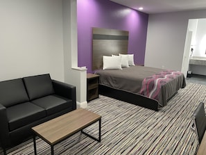 Una habitación de hotel moderna con una cama, un sofá negro, una mesa de centro de madera y una alfombra estampada.