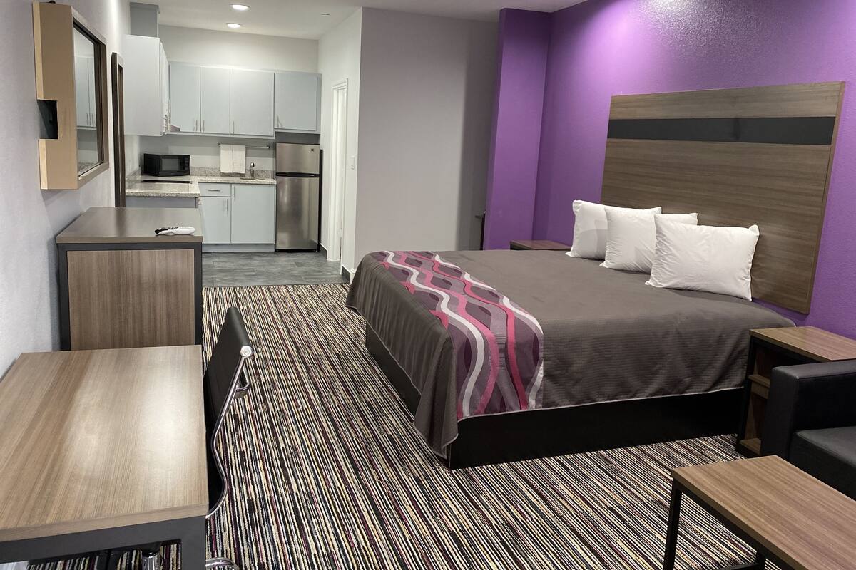 A hotel room with a bed, a desk, a chair, a small table, a microwave, and a refrigerator in the background.