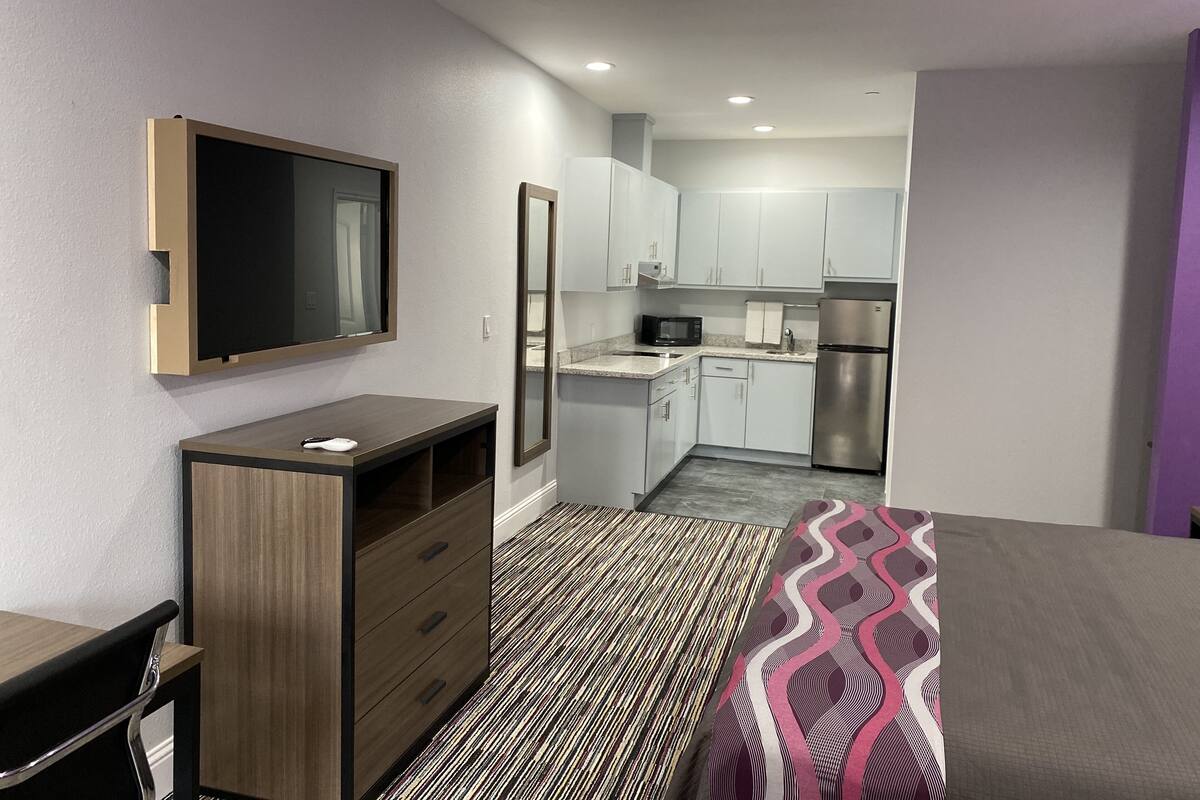 A compact hotel room with a bed, a desk, a dresser, a TV, a microwave, a refrigerator, and a kitchenette.