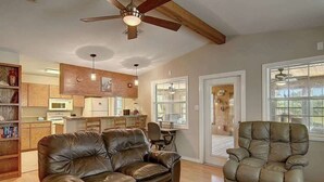 Un salon avec un canapé en cuir marron, un fauteuil inclinable, un ventilateur de plafond, un coin cuisine et un chien visible à travers une porte vitrée.