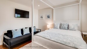 Ein modernes Schlafzimmer mit einem Himmelbett, einem dunklen Sofa mit gemusterten Kissen, einem flachbildfernseher an der Wand und zwei gerahmten Kunstwerken.