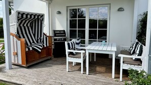 En overdækket terrasse med en træstol, et bord med stole og en grill.