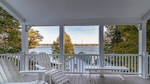 Een overdekt terras met witte Adirondack-stoelen en een tafel, met uitzicht op water en bomen.