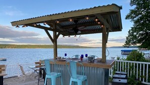 Uma área de bar ao ar livre com pérgola de madeira, dois bancos de bar azuis e uma mesa com cadeiras.