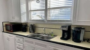 Een keuken met witte kasten, een zwart aanrecht, een spoelbak, een magnetron, een koffiezetapparaat en een raam met jaloezieën.