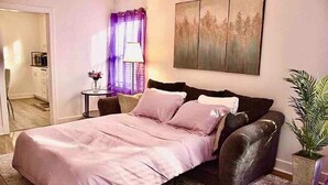 Een slaapkamer met een bed, een klein tafeltje met een lamp, een kamerplant en een schilderij aan de muur.