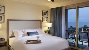 A hotel room with a large bed, a ceiling fan, a nightstand with a lamp, and a view of the ocean through a large window. - OCEAN FRONT, PRIVATE BEACH Luxury Suite with Balcony sleeping 8 adults (Hilton Head Island)