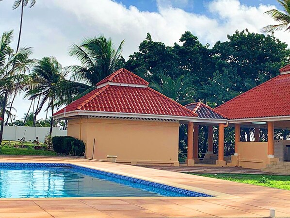 Una piscina con borde de baldosas, dos edificios con techos rojos y palmeras al fondo.
