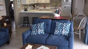 A living room with a blue sofa, a wicker coffee table, and a kitchen area with white cabinets and a refrigerator. - Pet friendly on the beach with private balcony! (Fort Walton Beach)