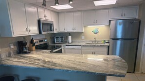 A kitchen with white cabinets, a stainless steel refrigerator, a microwave, a coffee maker, and a toaster. There are three stools at the island. - Pet friendly on the beach with private balcony! (Fort Walton Beach)