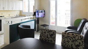 A compact living space with a kitchenette, a dining area with a table and chairs, a television, and a balcony with a view of buildings.