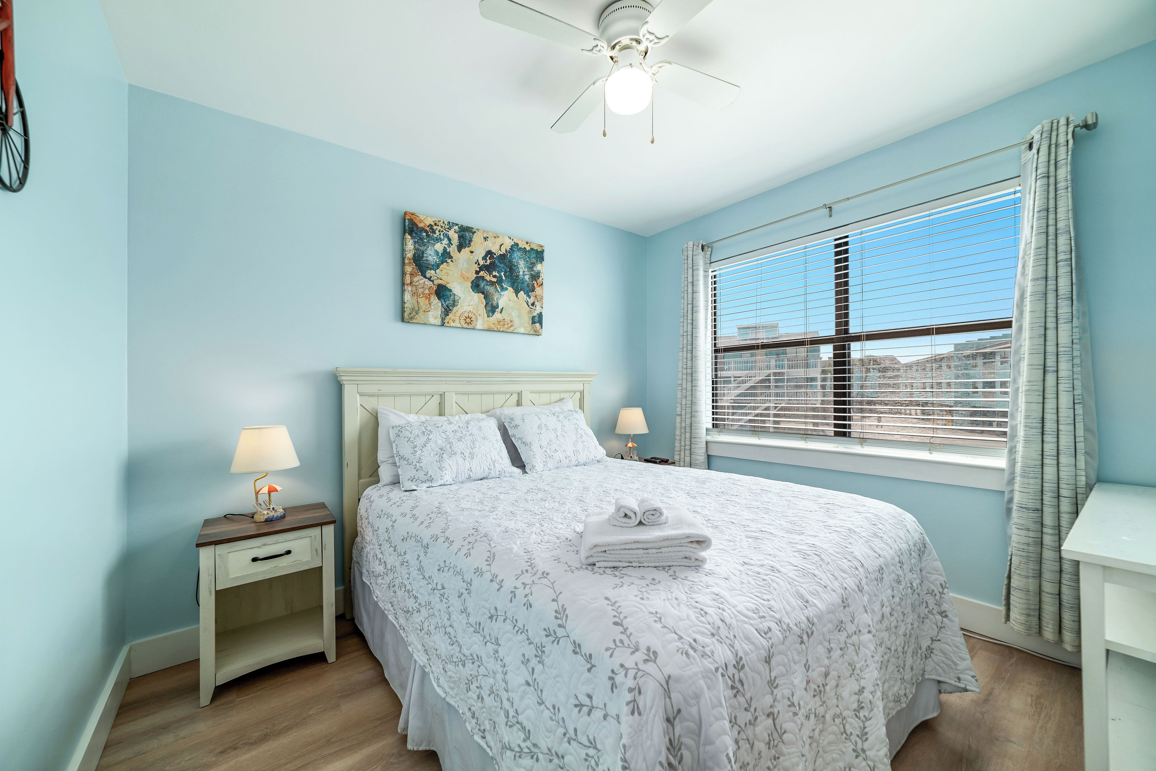 A bedroom with a bed, a nightstand, a lamp, a ceiling fan, a window with blinds, and a painting on the wall.