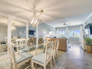 A spacious dining area with a glass table and white chairs, a kitchen with a large window, and a ceiling fan.