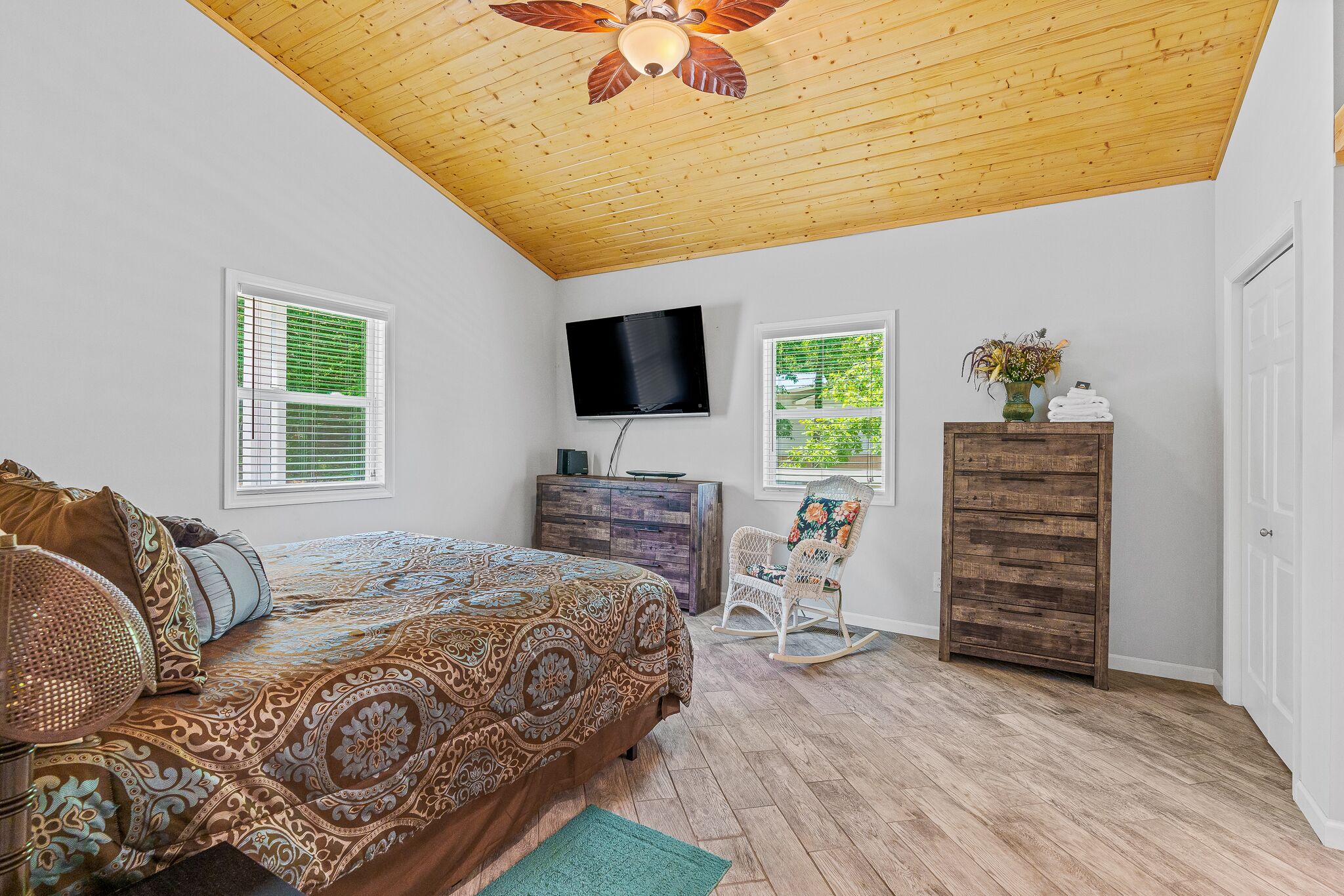 Une chambre avec un plafond en bois, un lit avec une literie à motifs, un téléviseur fixé au mur et une commode.