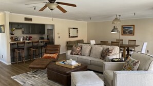 A spacious living room with a sofa, ottoman, and chairs, a dining area with a table and chairs, and a kitchen with a refrigerator and bar stools.