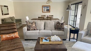 A living room with a sofa, ottoman, and chairs. There is a dining table with chairs and a picture on the wall.