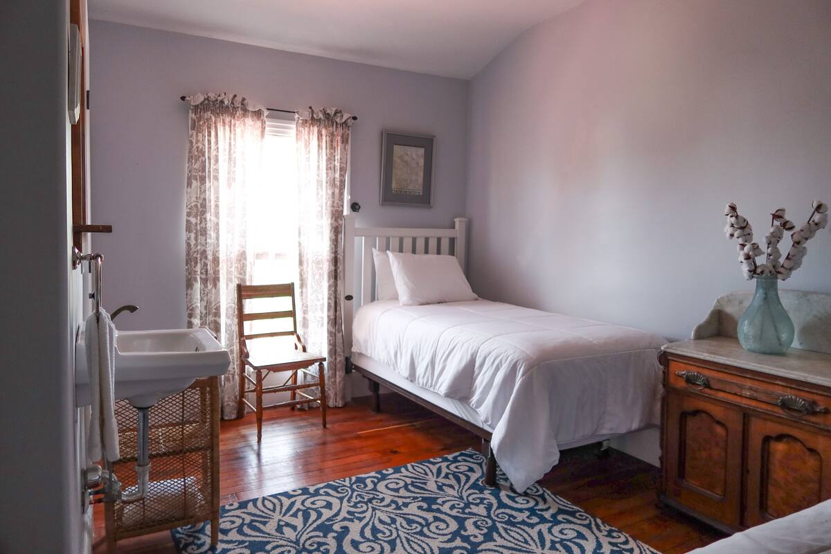 Ein Schlafzimmer mit einem Einzelbett, einer Holzkommode, einem weißen Nachttisch, einem Sessel, einem kleinen Waschbecken und einem gemusterten Teppich.
