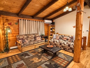 Ein gemütliches Blockhaus-Interieur mit Holzbalkendecke, zwei Sofas mit gemusterten Kissen, einem Couchtisch und einer dekorativen Wandleuchte.