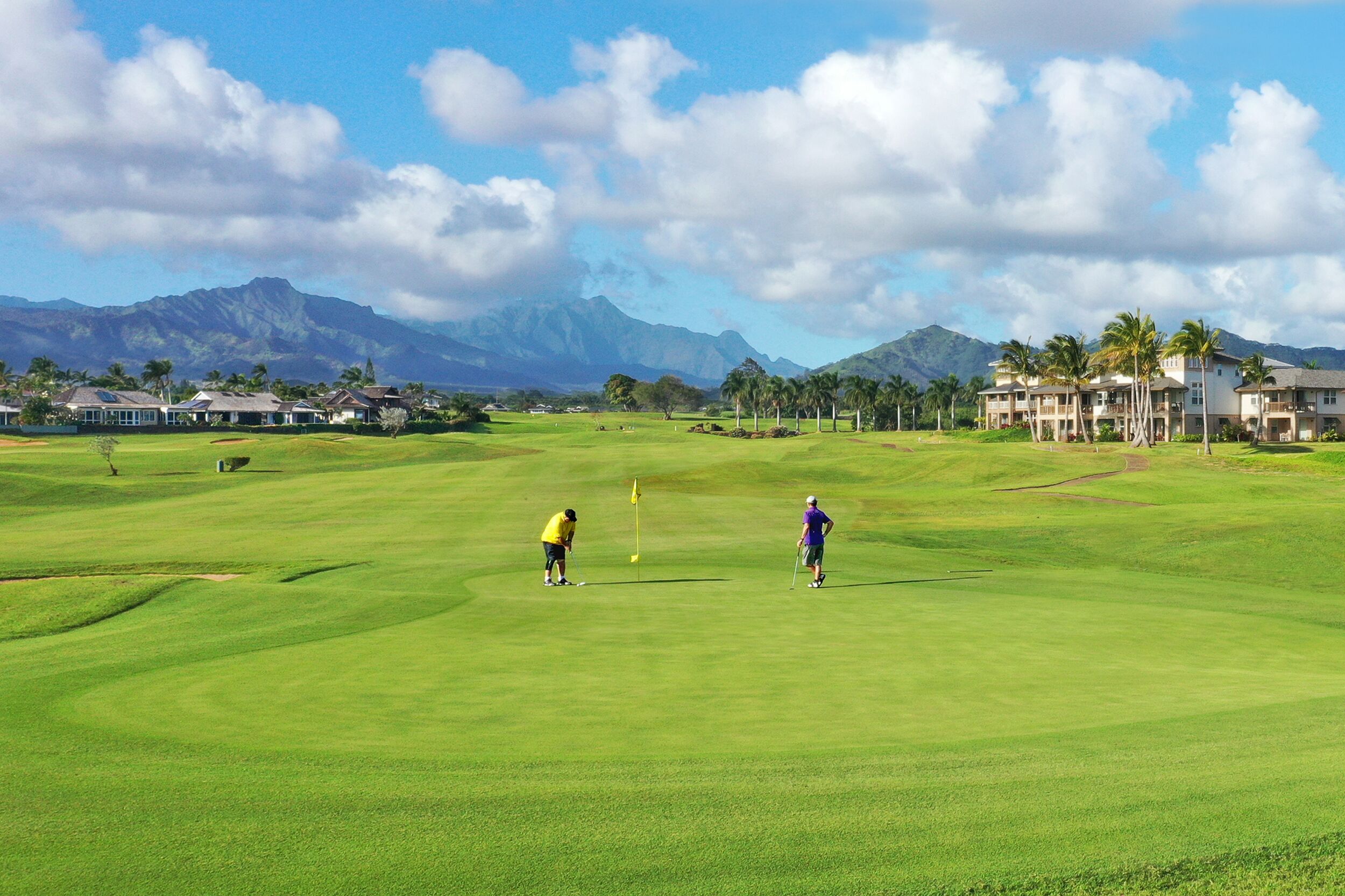 Un parcours de golf avec une chaîne de montagnes en arrière-plan, une pelouse bien entretenue et quelques bâtiments visibles au loin.