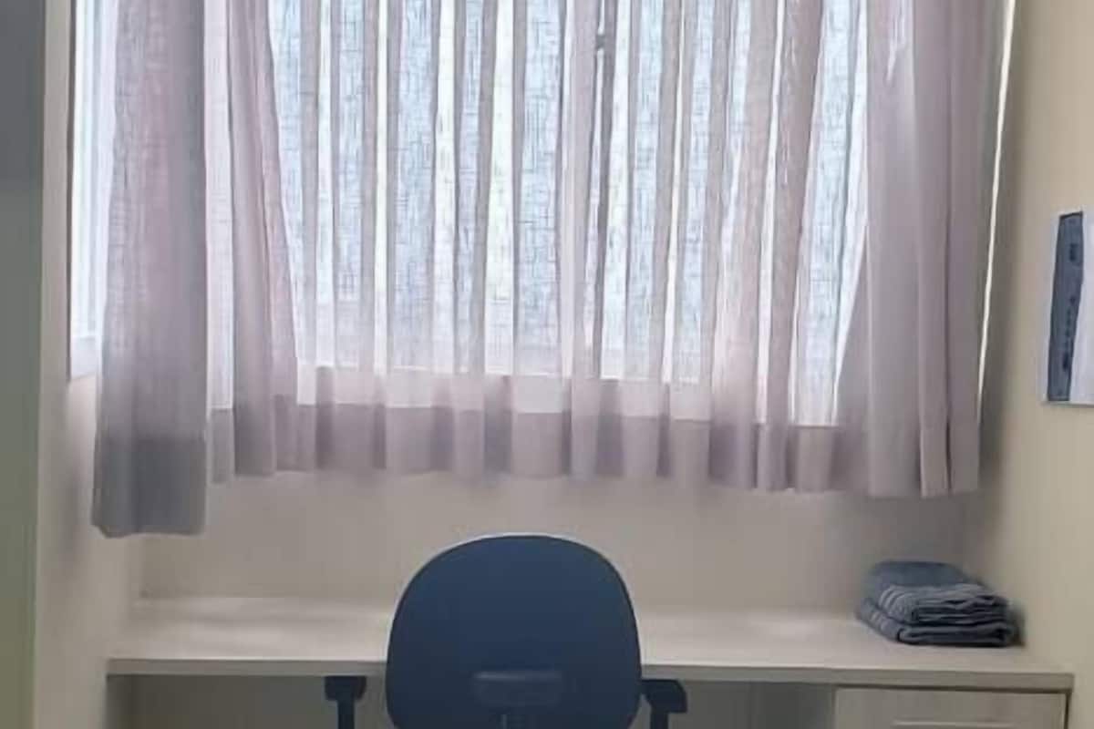 Ein Zimmer mit Schreibtisch, Stuhl und einem Fenster mit durchsichtigen Vorhängen.
