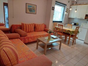 Ein Wohnzimmer mit einer Sofagarnitur und einem Esstisch mit Stühlen. Es gibt eine Küchenzeile mit Schränken und einem Kühlschrank.