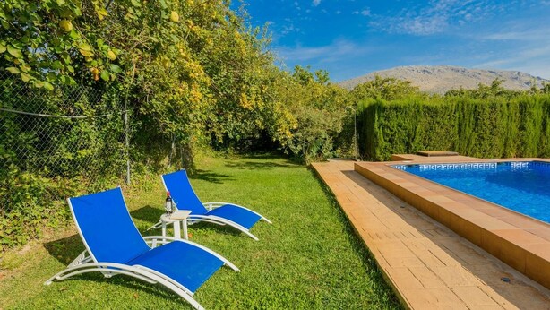 A swimming pool with a wooden deck, two blue lounge chairs, and a grassy area with trees and a fence.