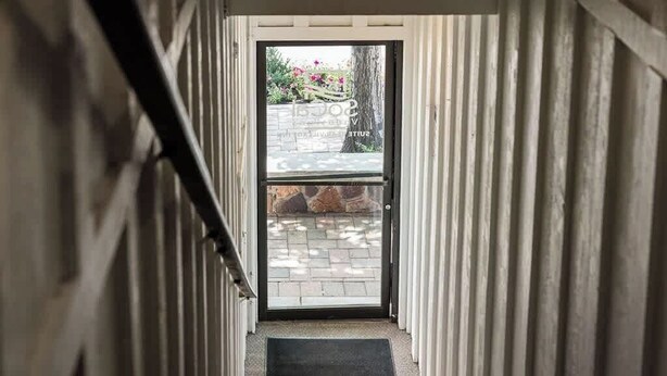 Ein schmaler Flur führt zu einer Tür, hinter der eine Steinwand und ein kleiner Garten zu sehen sind.