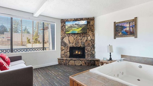 A room with a stone fireplace, a large window with blinds, a sofa, a bathtub, and artwork on the walls. - Teddy Bear, Perfect Location in the Village (Big Bear Lake)