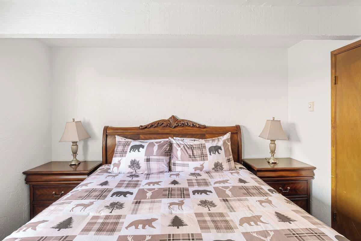 A neatly made bed with a patterned bedspread and two pillows, flanked by nightstands with lamps, in a room with white walls and a wooden door.