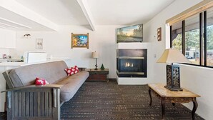 A modern living room with a fireplace, a sofa, a coffee table, and a view through a large window. - Grizzly Bear, Perfect Location in the Village (Big Bear Lake)