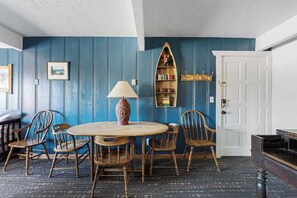Una habitación con una mesa redonda de madera, cuatro sillas de madera, una lámpara, una estantería y una puerta blanca.
