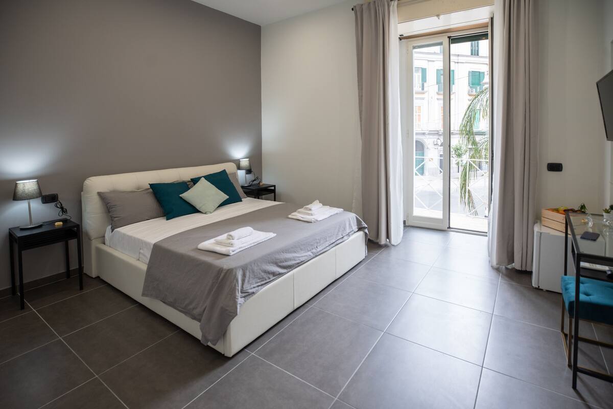 A modern hotel room with a large bed, a desk, and a view of the street through the window.