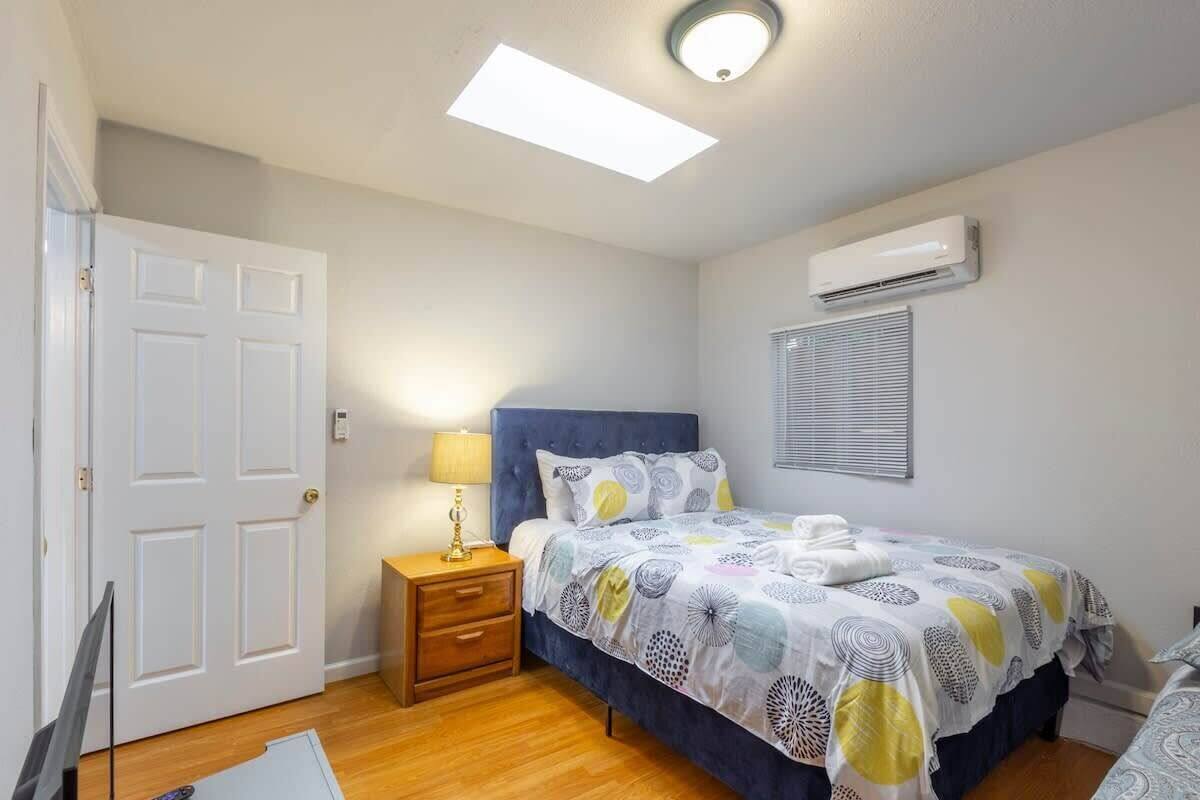 A bedroom with a bed, bedside table, lamp, ceiling light, air conditioning unit, and a door.