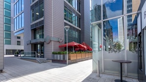 A modern building with a glass facade, outdoor seating area with red umbrellas, and a small table.