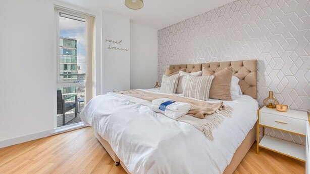 A modern bedroom with a large bed, a view of a cityscape through a sliding door, and a geometric-patterned wall.