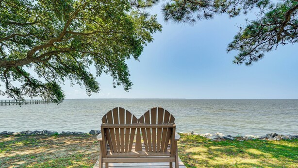 A wooden Adirondack chair on a wooden deck by a lake with a dock in the background. - Waterfront Nags Head Townhome w/ Amenity Access! (Nags Head)
