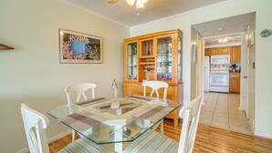 A dining area with a glass table, white chairs, a ceiling fan, and a kitchen visible through an open doorway. - Waterfront Nags Head Townhome w/ Amenity Access! (Nags Head)