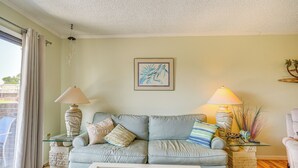 A living room with a light blue sofa, two side tables with lamps, a framed artwork on the wall, and a view through a large window. - Waterfront Nags Head Townhome w/ Amenity Access! (Nags Head)