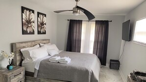 Um quarto bem organizado com uma cama, cabeceira de madeira, um ventilador de teto, uma mesinha de cabeceira com um vaso de flores, um aparador e uma TV montada na parede.