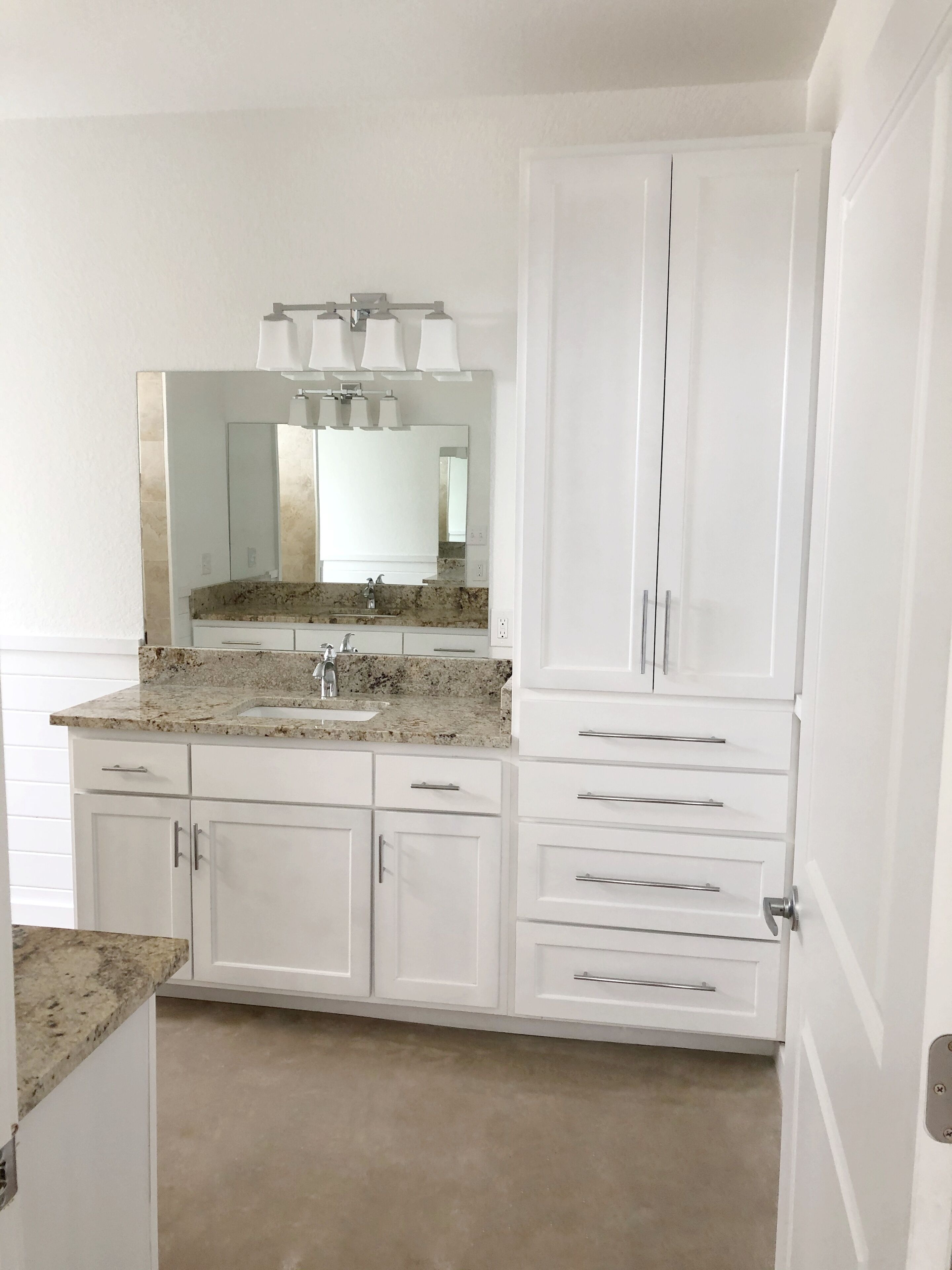 A modern bathroom with a granite countertop, a large mirror, white cabinetry, and a shower area.
